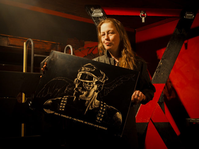 Sahra Shaubach in the room she and volunteers extensively rehabbed inside the basement of the 225 E. 5th Ave property, holding a poster from the The Eagle, a renown Baltimore leather bar. (Photo by Zachariah Hughes, KSKA)