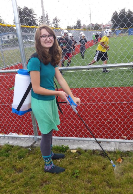 Anna McDonald sprays a patch of grass as the Kodiak football team practices in the background. (Photo by Kayla Desroches/ KMXT)