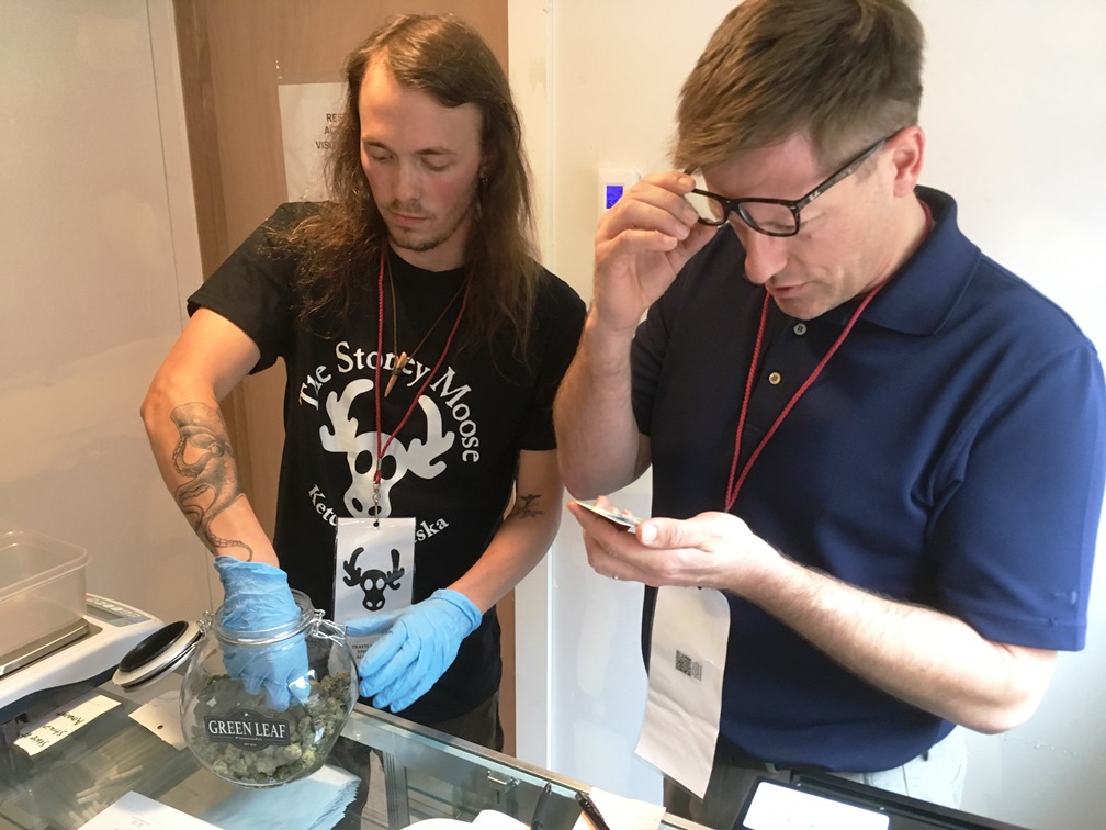 Travis Carter gets some cannabis buds out to measure for a customer while Mark Woodward checks an ID on the opening day for Ketchikan’s Stoney Moose. (Photo by Leila Kheiry/KRBD)