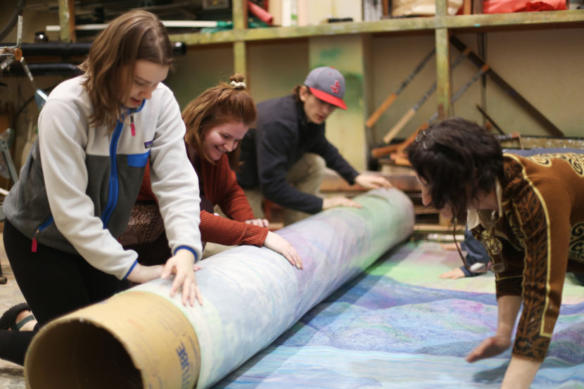JDHS Art Club members Riley Stadt, Janessa Goodman, and Kane Ginter roll up the 2018 Alaska Folk Festival backdrop with adviser Heather Ridgway. (Photo by Annie Bartholomew/KTOO)
