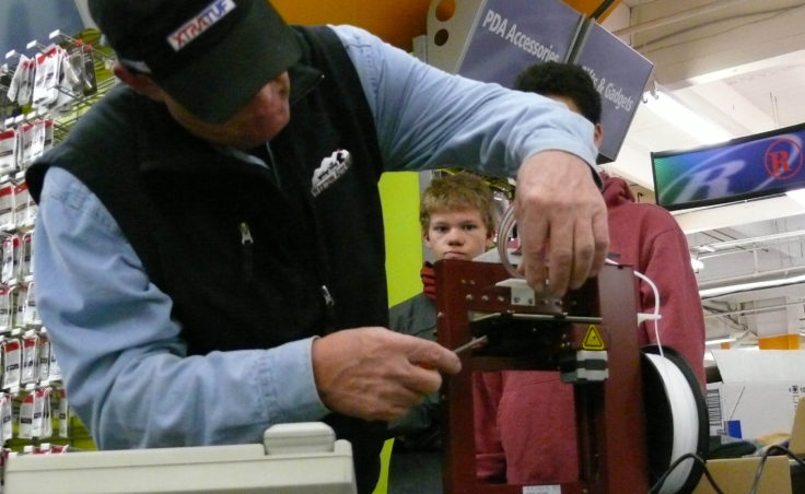 John Weedman adjusts a component of the 3D printer as TMHS freshman William Wallace looks on. (Photo by Jeremy Hsieh/ KTOO)