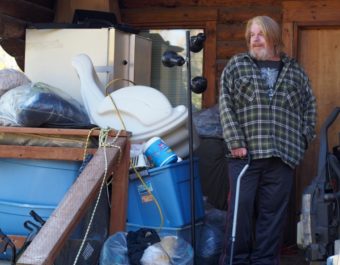 Scott Harrison standing outside the cabin he rents in Big Lake. It’s too small inside for him to fit many of his possessions, so he keeps more durable items like clothes and dry-goods outside. (Photo by Zachariah Hughes/KSKA)