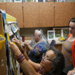 Della Cheney, left, and Clarissa Rizal work on braiding the side borders for the Weavers Across Waters Chilkat/Ravenstail community robe on Monday, August 22, 2016, at the Sealaska Heritage Institute, Juneau. One of three of Rizal's daughters, Lily Hope watches the weavers work on the robe, which will be part of the Huna Tribal House opening celebration. Hope is also an accomplished weaver, actress and storyteller. (Photo by Tripp J Crouse/KTOO)