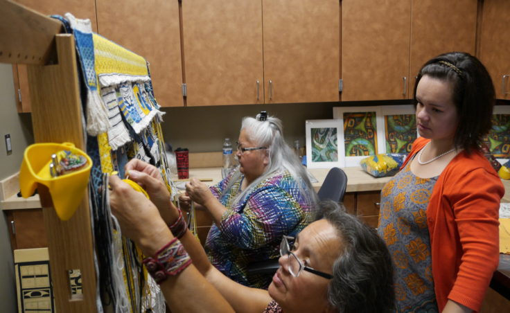 Della Cheney, left, and Clarissa Rizal work on braiding the side borders for the Weavers Across Waters Chilkat/Ravenstail community robe on Monday, August 22, 2016, at the Sealaska Heritage Institute, Juneau. One of three of Rizal's daughters, Lily Hope watches the weavers work on the robe, which will be part of the Huna Tribal House opening celebration. Hope is also an accomplished weaver, actress and storyteller. (Photo by Tripp J Crouse/KTOO)
