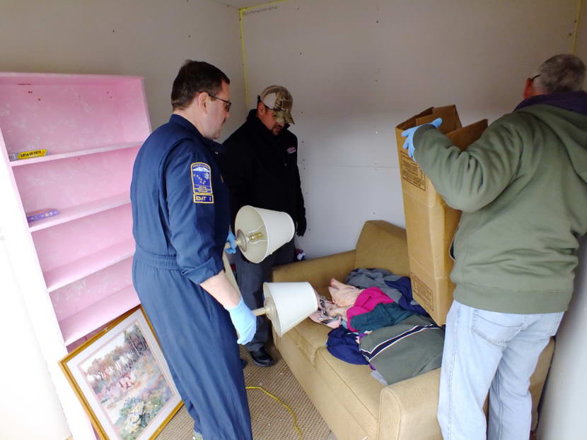 Instructors Ernie Misewicz, Jason Mardirosian, and Don Cuthbert position furnishings and appliances a recent fire investigator training exercise at Juneau’s Hagevig Regional Fire Training Center. A dead pig has been dressed in clothes and covered with a blanket on the couch to mimic a human fatality. (Photo by Matt Miller/KTOO)