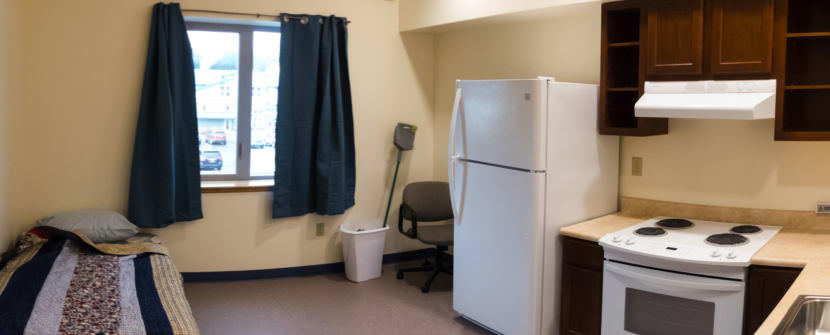 An efficiency apartment with a small bed with a quilt on it, a window,, a full sized refrigerator, and a small kitchenette area