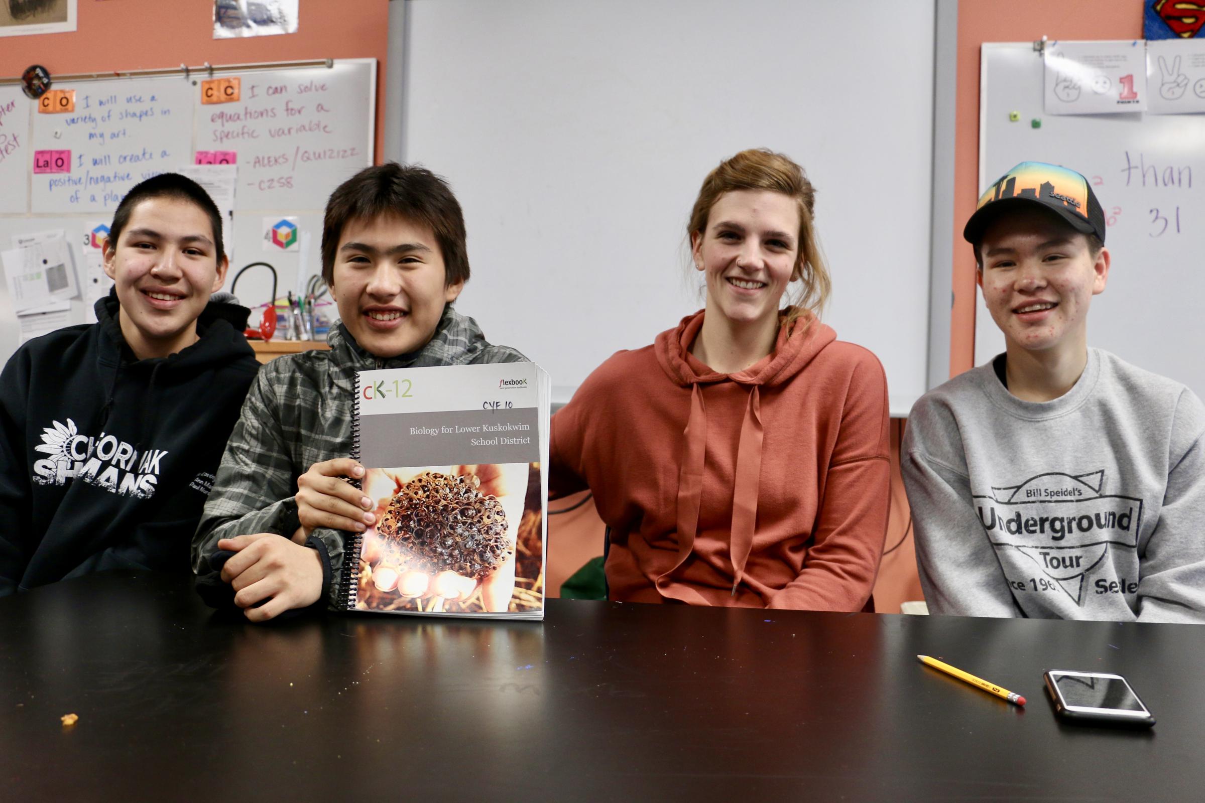 Chefornak High School students Matthew Erik, left, Clayton Panruk, pose with biology teacher Theresa Schallhorn, and fellow student Dawson Erik, with their new CK-12 Foundation biology textbook on December 16, 2017. (Photo by Christine Trudeau/KYUK)