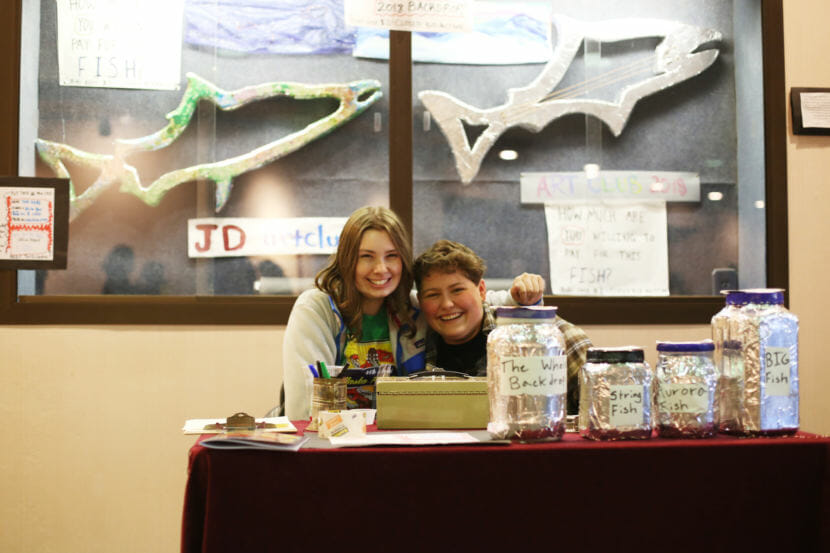 Two people sit at a table at the Alaska Folk Festival in front of fish sculptures. Juneau-Douglas High School Art Club co-President Riley Stadt and Secretary Theo Houck auction off fish sculptures to raise money for the club's trip to Region V Art Fest. (Photo by Annie Bartholomew/KTOO)