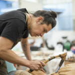 Lead carver Yéil Ya-Tseen Nicholas Galanin of Sitka uses an adze to carve the 40 foot T’aaku Kwáan Yanyeidí Healing kootéeyaa totem pole at Harborview Elementary School on 29, 2018. (Photo by Annie Bartholomew/KTOO)