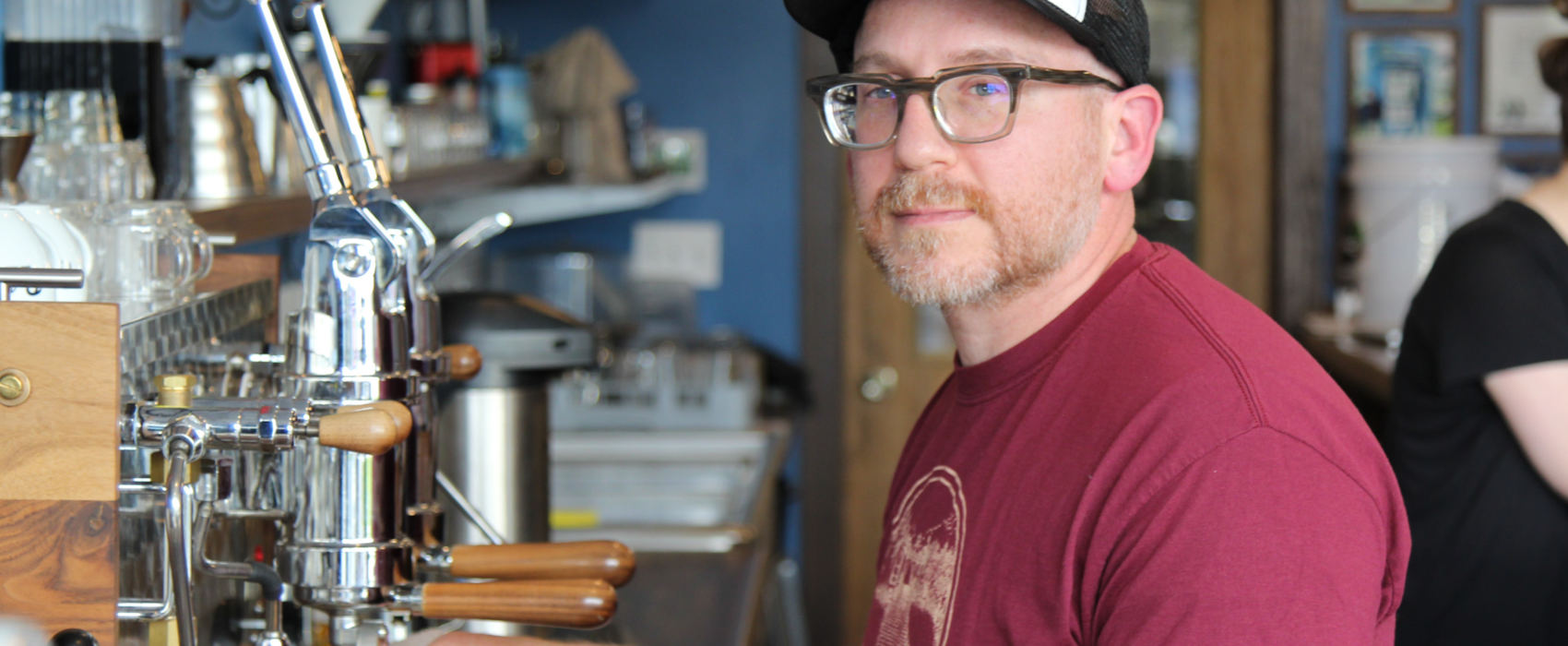 Austin Schwartz, with a short beard, glasses and a baseball cap, holds a cup of coffee under the nozzle of an espresso machine