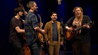 An image of a band surrounding a microphone with men playing upright bass, hollowbody electric guitar, singing and acoustic guitar. Screen capture of the Seattle-based band Revilla performing a Red Carpet Concert at KTOO during the 2018 Alaska Folk Festival featuring Alex Nelson, Alejandro Chavarria and Patrick Troll.