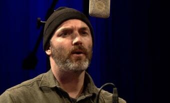 An image of a man in a black hat singing into a microphone. A video still of Seattle songwriter and guitarist Eli West performing a Red Carpet Concert at KTOO Public Media during the 2018 Alaska Folk Festival in Juneau.