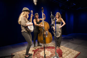 Three women stand around a microphone playing upright bass, washboard and banjo. Alaska band Damn Fantasies and the Could Be Possibilities perform a Red Carpet Concert at KTOO Public Media on April 12, 2018. (Photo by Annie Bartholomew/KTOO)
