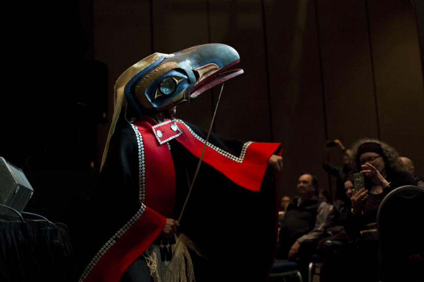 A man in a button blanket and raven mask looks into light at Centennial Hall. Gene Tagaban dances with Khu.éex' at Centennial Hall on Monday, January 28, 2019. Tagaban played flute and sang as part of the performance. (Photo by Annie Bartholomew/KTOO)