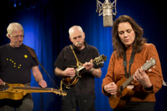 A woman plays ukulele into a microphone, while accompanied by a man on a dobro and another gentleman on mandolin. American composer and lyricist Georgia Stitt performs "The Things That Are True" as Snow Children during the 2019 Alaska Folk Festival as part of KTOO Public Media's Red Carpet Concert series. The group included co-composer Bob Banghart on Mandolin and Eric Graves on dobro. (Photo by Annie Bartholomew/KTOO)