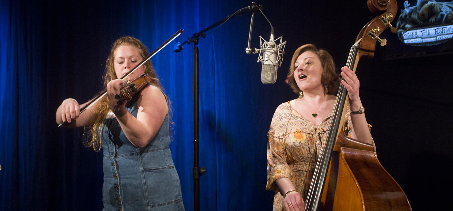 A woman plays violin while another woman playing upright bass sings into a microphone in a television studio.Katie Klan and Angela Brock of the Homer band Burnt Down House perform a Red Carpet Concert at KTOO Public Media during the 2019 Alaska Folk Festival. (Photo by Annie Bartholomew/KTOO)