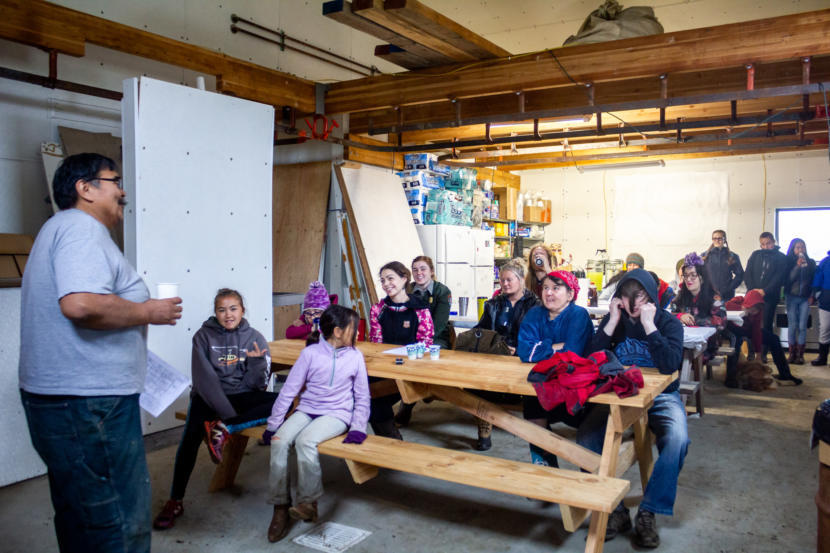 Man stands before group of visitors seated at picnic tables inside large building.