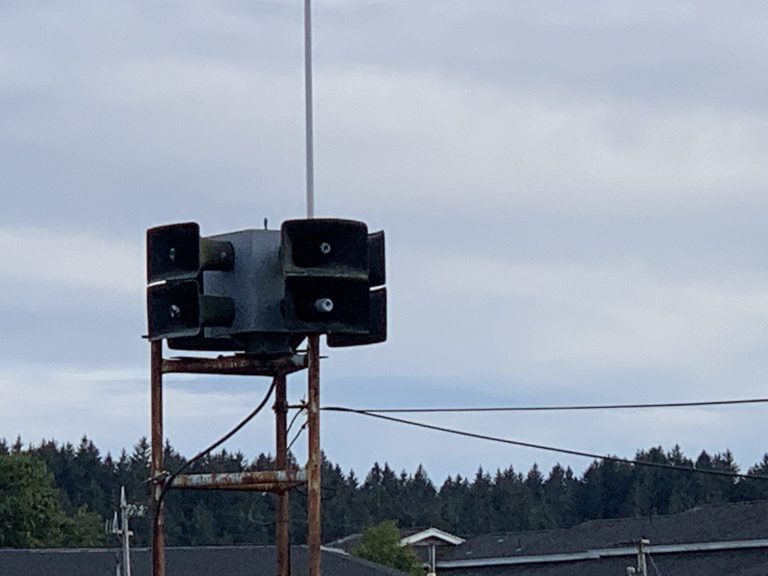 The Kodiak tsunami siren photographed with the sky above it and a tree line below