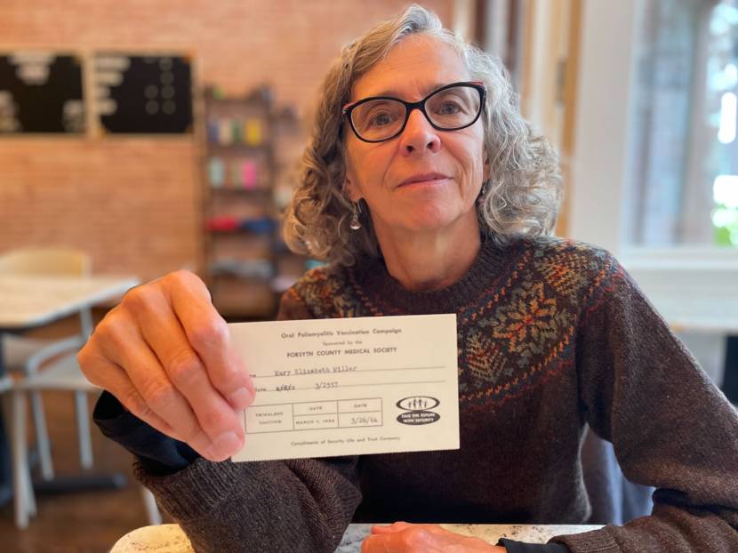 A seated woman holding up a polio vaccine card for the camera