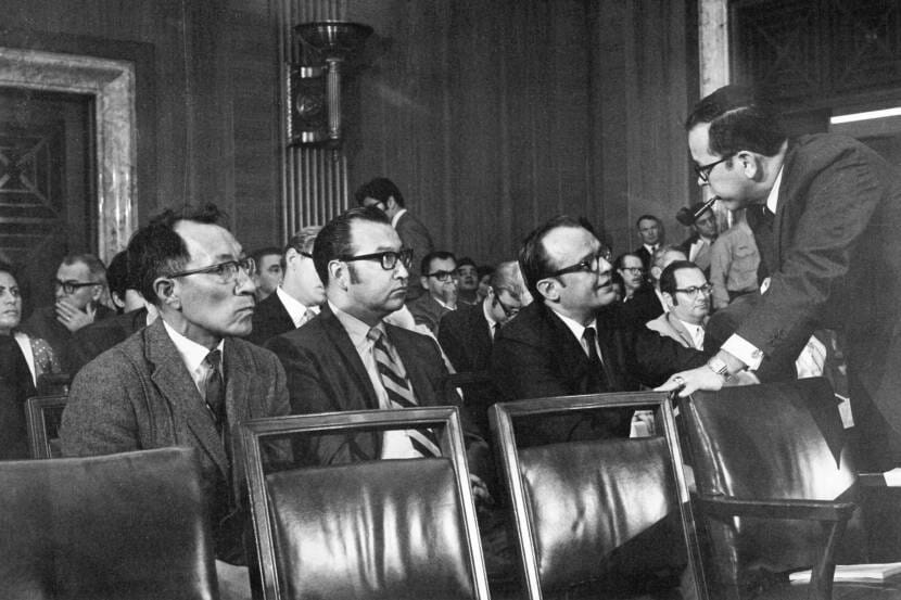 A black-and-white photo of a man with a pipe, standing, speaking to three seated men in 