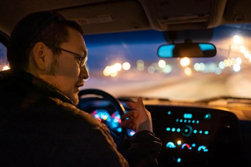 A photo taken from the back seat of a car of a man driving at night on snowy streets