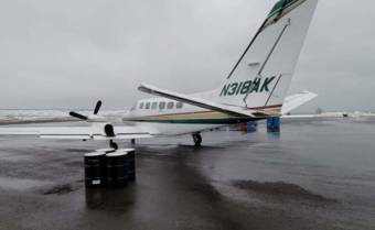 A twin engine plane sitting on a wet runway next to four fuel drums