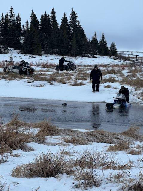 Several people looking at a snowmachine half-sunk in a frozen river