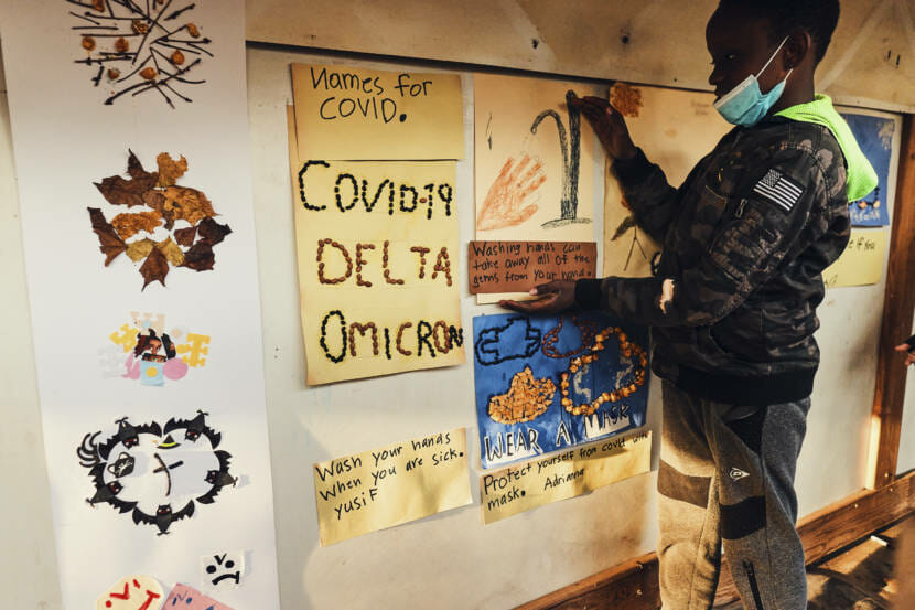 A woman standing by a wall covered with children's art work about covid and covid safety