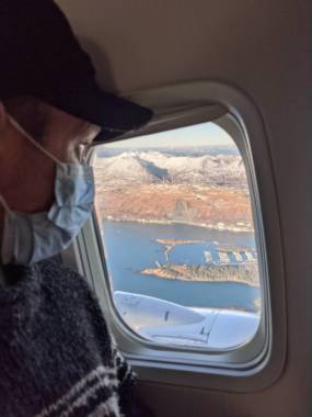A man in a surgical mask looking out an airplane window at a harbor