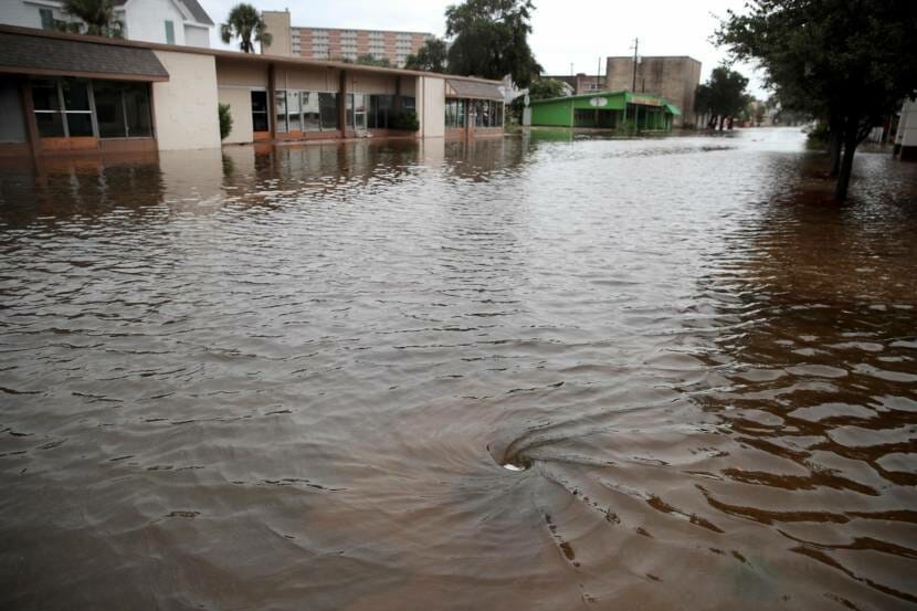 A flooded street with a small whirlpool where muddy water is going down a sewer drain
