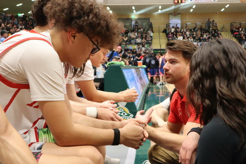 A basketball team sits courtside talking to their coach in a crowded arena
