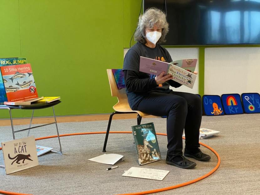 A woman in a mask reads aloud from a book in a library 