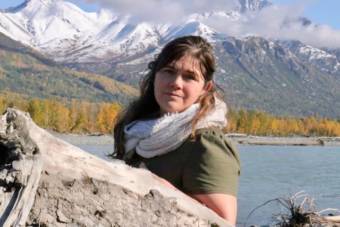 A portrait of a woman standing outside by a river, with snowy mountains behind her