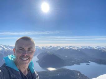 A selfie taken by a man standing on a mountain with bays and snowy mountains in the background