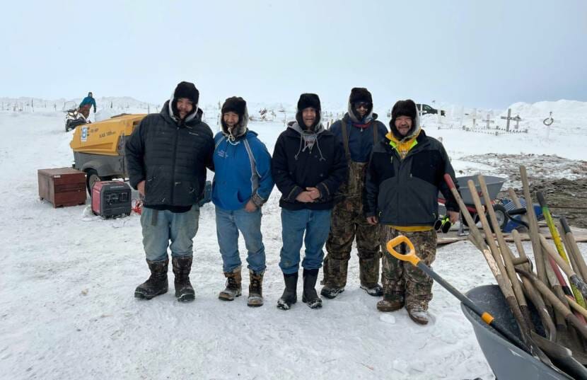 Five men stand at the edge of a snowy graveyard, with a wheelbarrow filled with shovels beside them