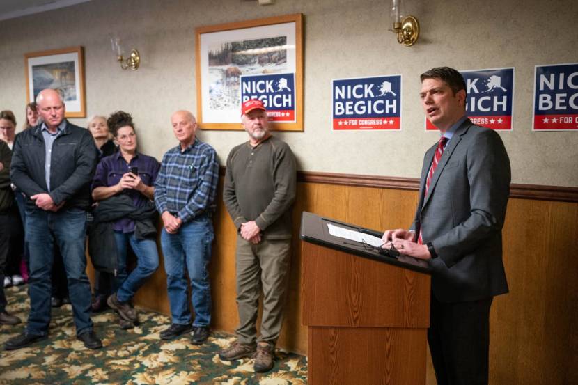 Nick Begich, wearing a grey suit, speaks from behind a lectern with several Nick Begich campaign posters on the wall behind him