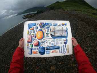Hands holding a drawing of objects found on a beach in front of an actual beach