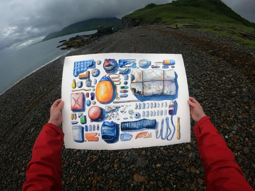 Hands holding a drawing of objects found on a beach in front of an actual beach