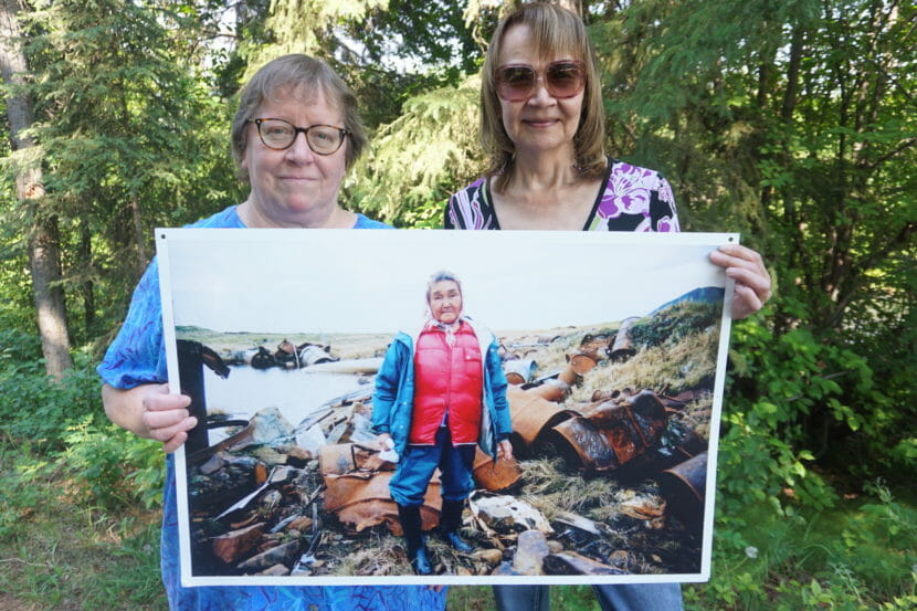 Two women, standing outside, hold a large photo of another woman