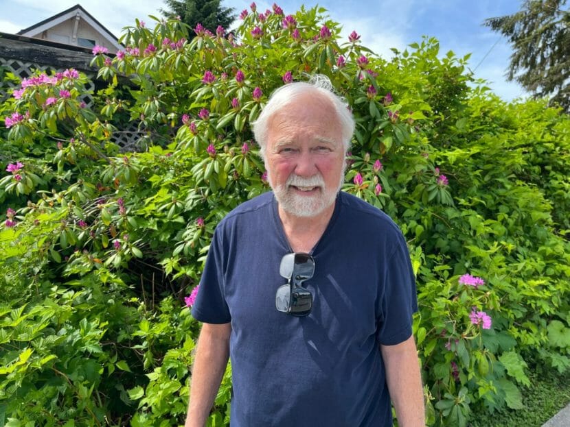 An older man standing in front of a shrub with two pairs of glasses hanging from his t-shirt collar