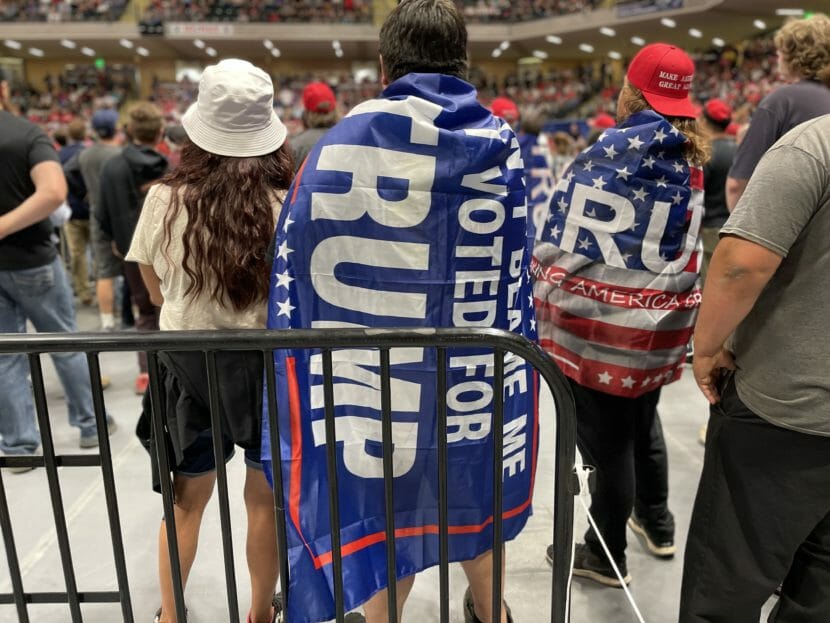 A person stands in the middle of a crowded arena with a flag draped over their shoulders that reads "Don't blame me. I voted for Trump."