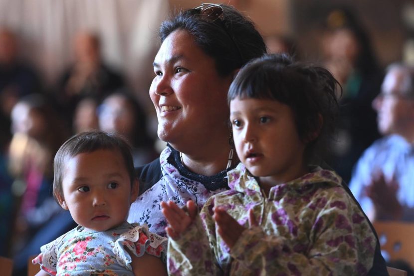 A smiling woman holds two small children, who are clapping