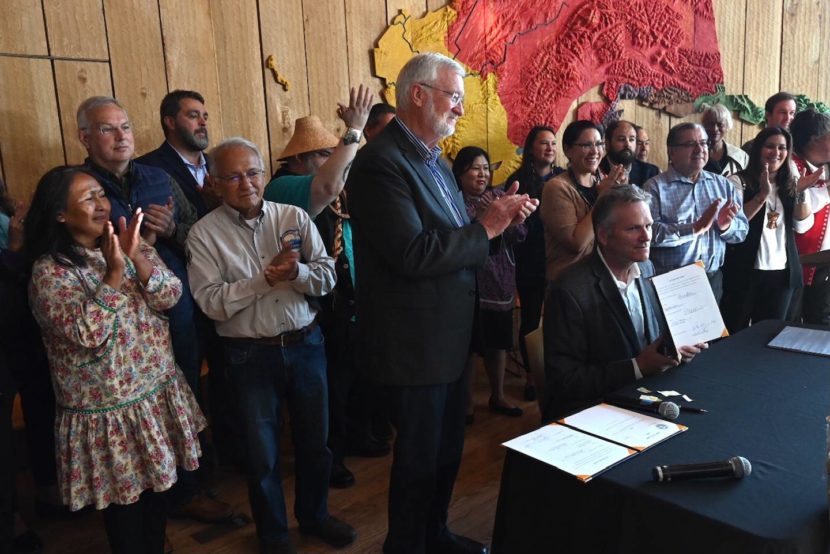 The governor seated at a desk, holding up a signed bill, while people stand around him and clap