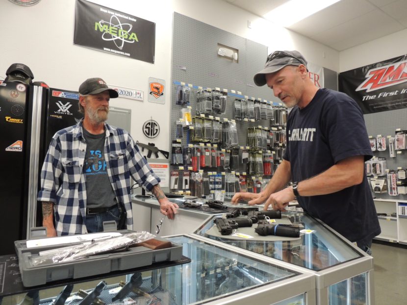 Two men on either side of a glass display case that has several pistols laid out on top of it