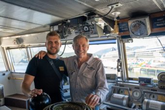 Two men stand on the bridge of a ferry with their arms around each others' shoulders