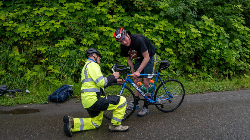 Man holds up a racing bicycle while a volunteer in a high viz suit kneels in front of the handlebars to fix something