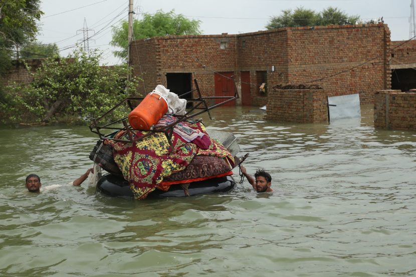 Two men, up to their necks in water, guiding a raft stacked with possessions away from a flooded brick building