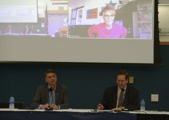 Two men sit at a table with Sarah Palin on a large screen behind them