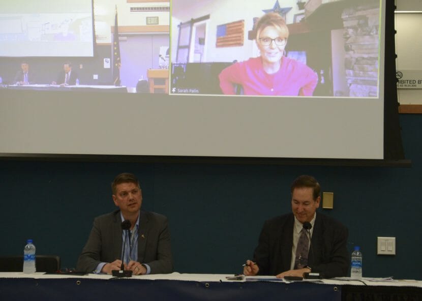 Two men sit at a table with Sarah Palin on a large screen behind them