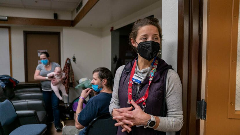 A woman wearing a mask stands in a room with her fingers interlaced. Behind her, a man sits while a woman carries a baby.